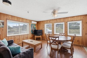 A room with wooden walls, a ceiling fan, a television on a stand, a dining table with chairs, and a sofa with cushions.