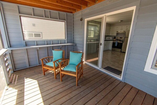 A covered deck with wooden flooring, two wicker chairs with teal cushions, and a sliding glass door leading to a kitchen area.