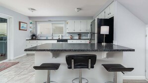 A modern kitchen with a granite countertop, white cabinets, and a black refrigerator. There are three bar stools and a window with blinds. - #155 Nestucca Sunrise · Riverfront, Close to Ocean Beaches, Large Yard (Pacific CIty)