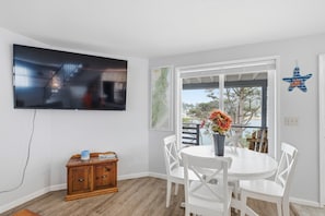Una habitación con televisión de pantalla plana en la pared, una mesa de comedor con sillas, un armario de madera y una ventana con vista a árboles y una valla.