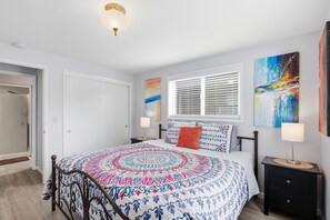 A bedroom with a bed, bedside tables, a window with blinds, and artwork on the walls.