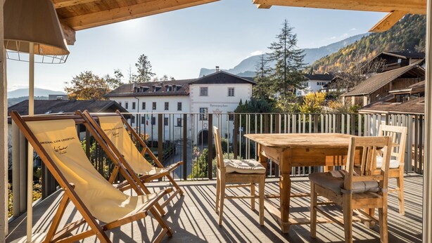 A wooden deck with a table and chairs, overlooking a village with traditional buildings and mountains in the background. - Apartment "Berg und Tal - Schlosserhaus 31" with Balcony and Mountain View (Tisens Südtirol)