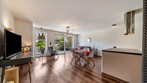 Una sala de estar moderna con un televisor de pantalla plana, piso de madera, una zona de comedor con una mesa y sillas rojas, y una cocina con encimera blanca y lámparas colgantes.