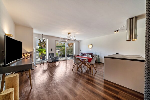 Un salon moderne doté d’un téléviseur à écran plat, de planchers en bois, d’un coin repas avec une table et des chaises rouges, et d’un espace cuisine avec un comptoir blanc et des luminaires suspendus.