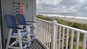Une véranda avec des fauteuils Adirondack bleus, une balustrade blanche et une vue sur la plage, où l’on aperçoit des parasols et des gens.