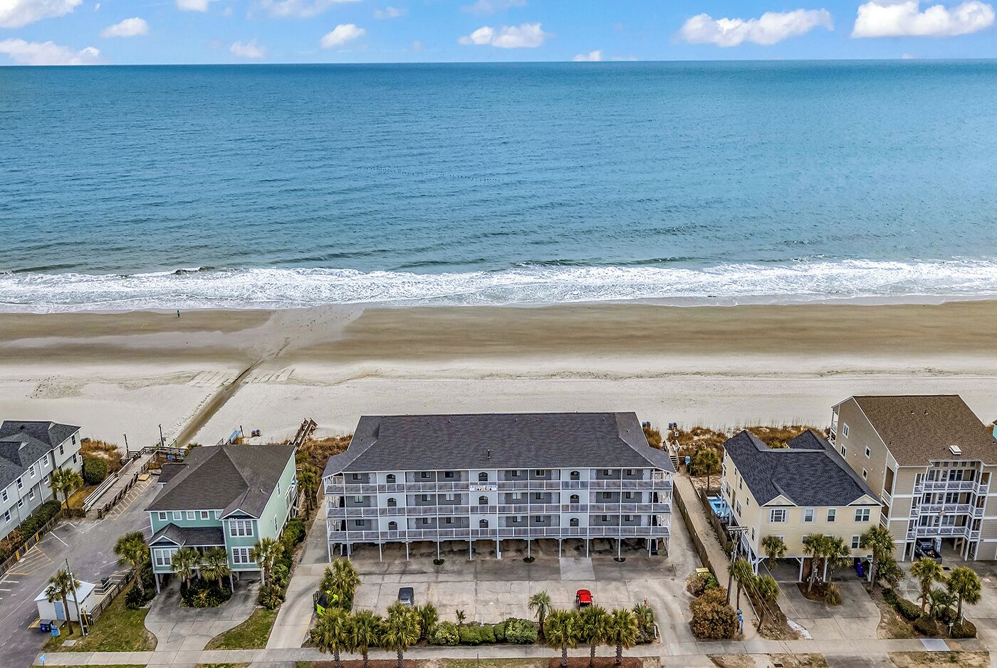 Un hotel frente a la playa con varias construcciones, un estacionamiento y vista al océano.