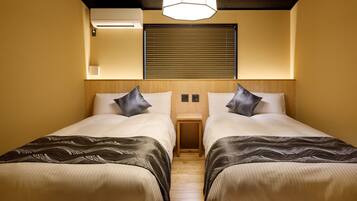A hotel room with two beds, each with a patterned bedspread, a bedside table, a wall-mounted air conditioner, and a window with blinds.