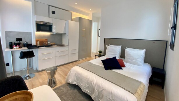 Quarto de hotel moderno com uma cama, pequeno espaço de cozinha, área de estar e vista para a cidade.