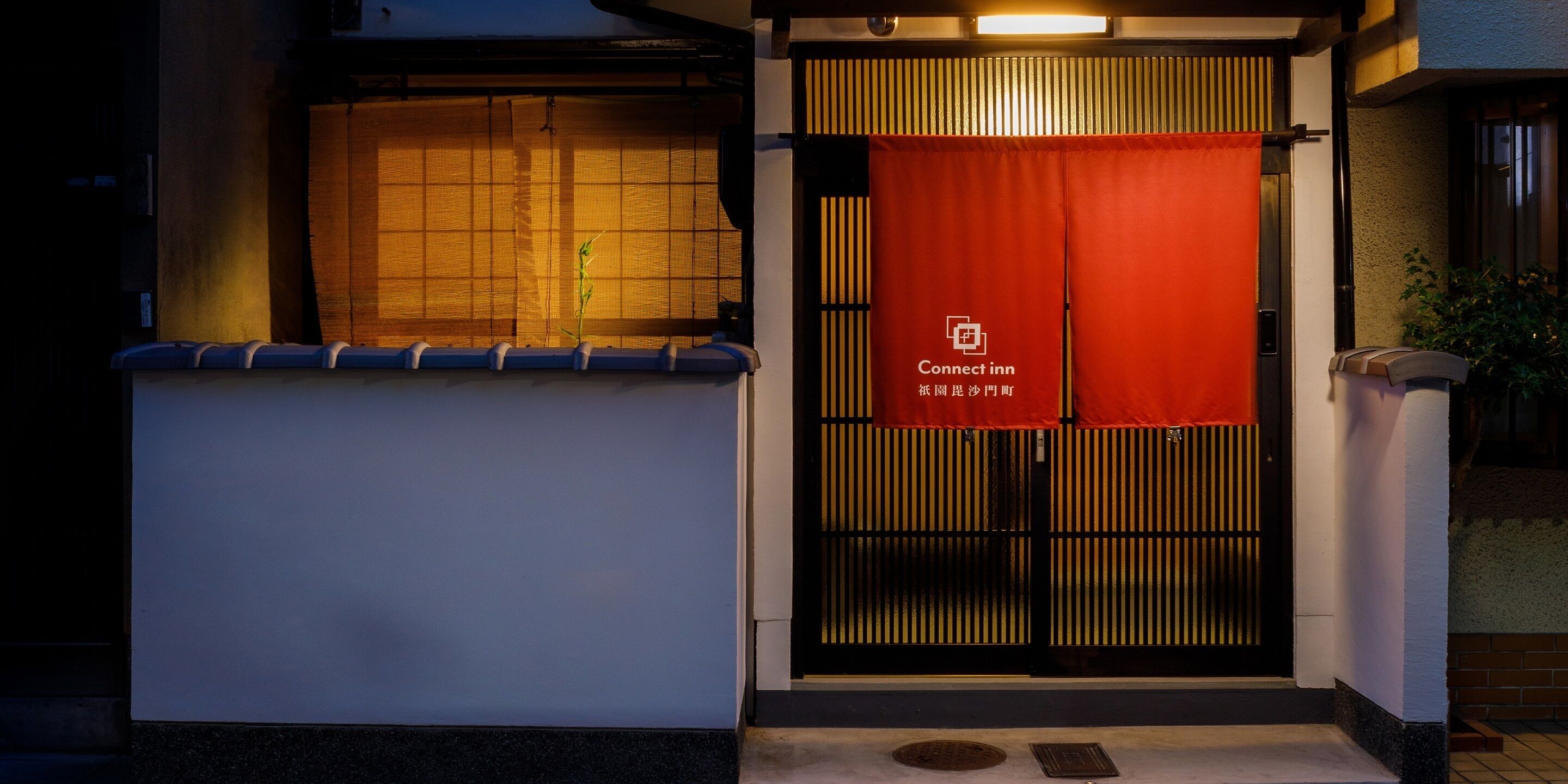 A hotel entrance with a traditional Japanese gate, a red banner with the hotel name, and a small plant in the window.