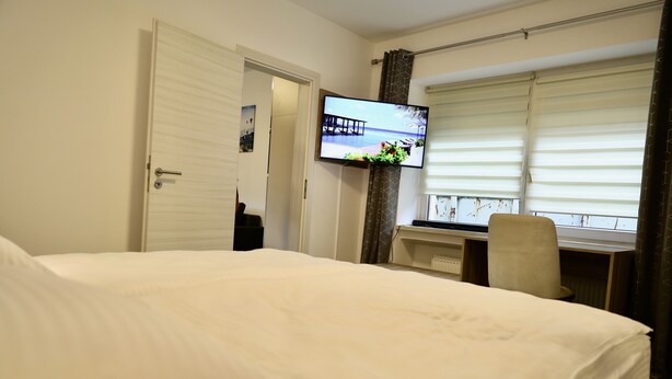 Ein Hotelzimmer mit einem großen Bett, einem Schreibtisch mit Stuhl, einem Wandfernseher und einem Fenster mit Jalousien.