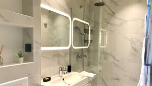 Un baño moderno con ducha de vidrio, tocador blanco con espejo y una pequeña planta en maceta.