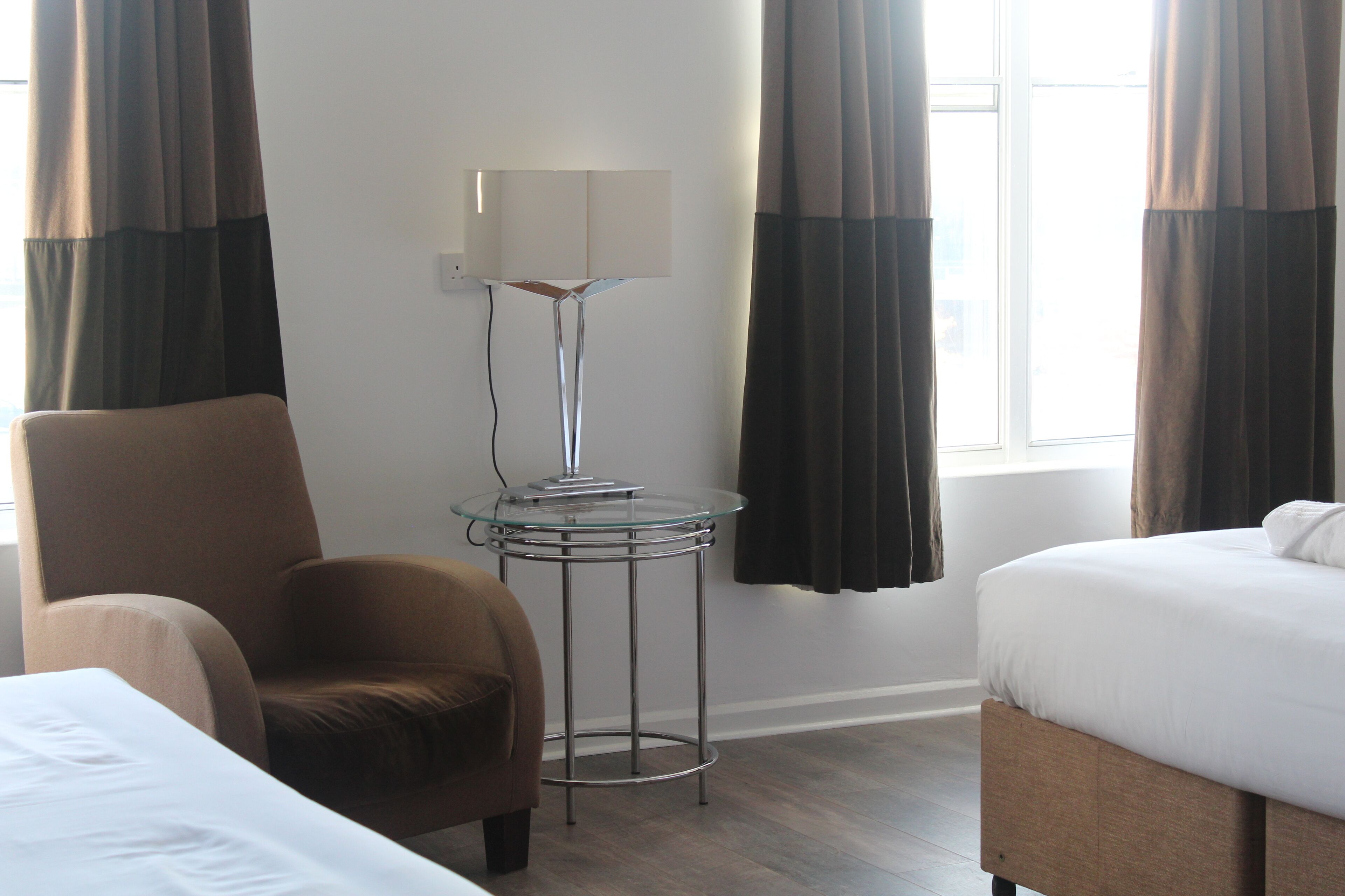 A hotel room with a bed, a chair, a small table with a lamp, and a window with curtains.