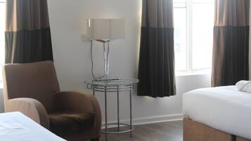A hotel room with a bed, a chair, a small table with a lamp, and a window with curtains.