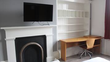 Een kamer met een open haard, een televisie erboven bevestigd, een bureau met stoel en ingebouwde planken.