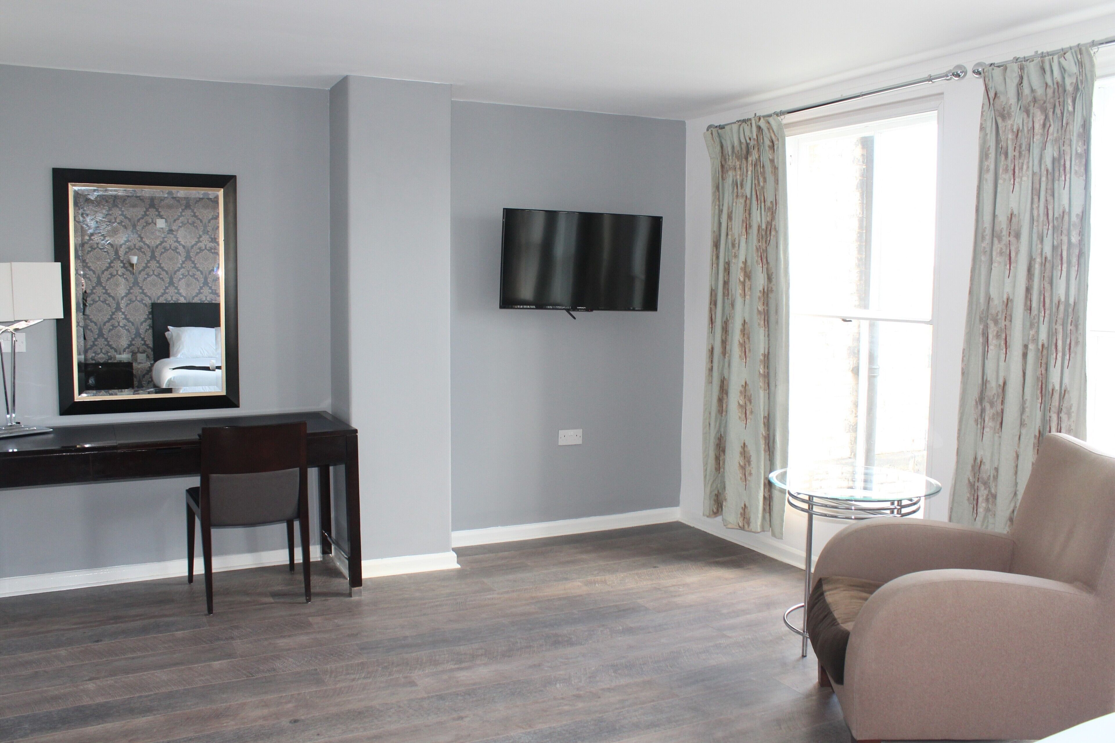 Una habitación de hotel moderna con una pared gris, un televisor de pantalla plana montado sobre un escritorio, una silla, una mesa redonda de vidrio y un ventanal con cortinas florales.