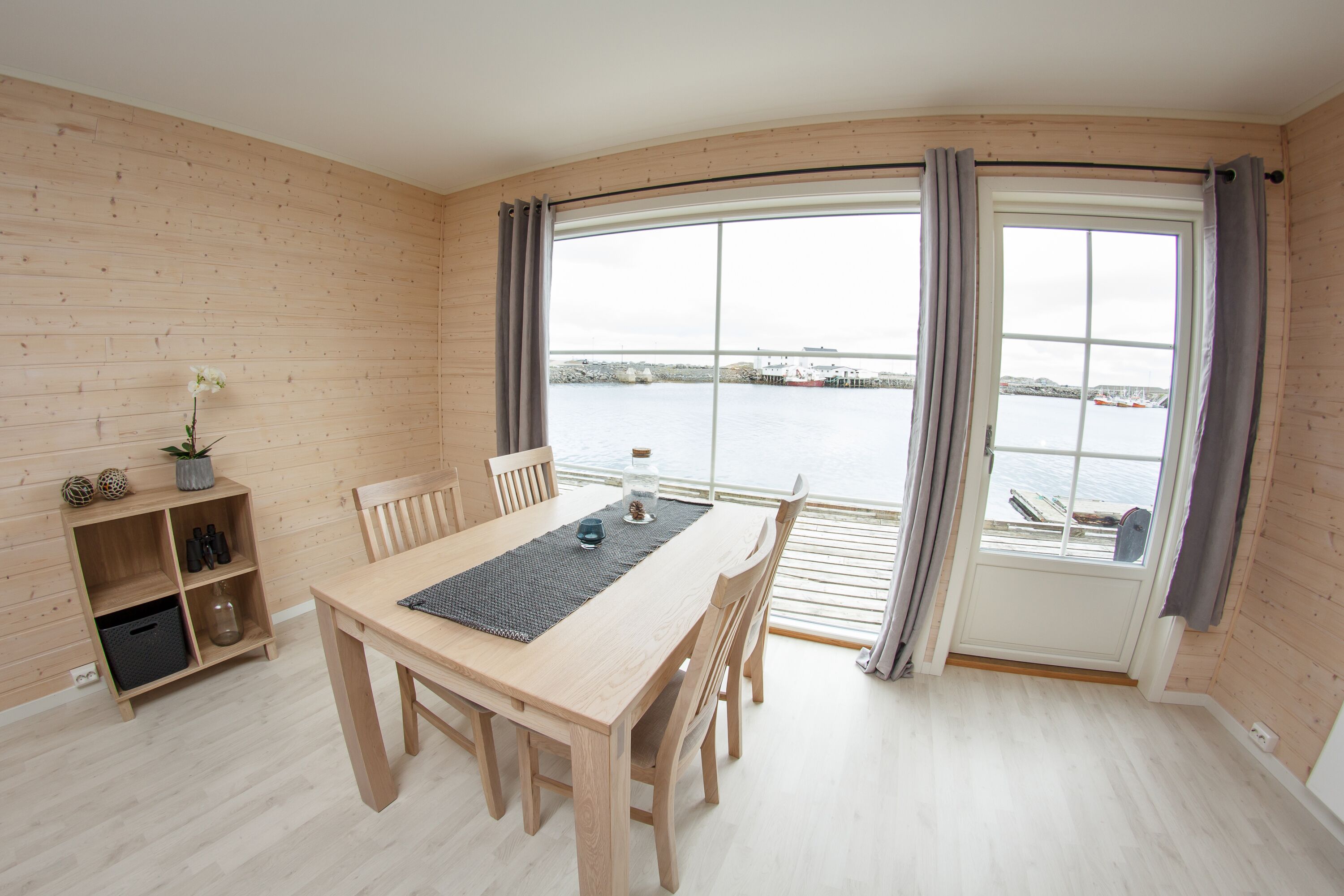 Un comedor de madera con una mesa, sillas y vistas al agua a través de amplios ventanales.
