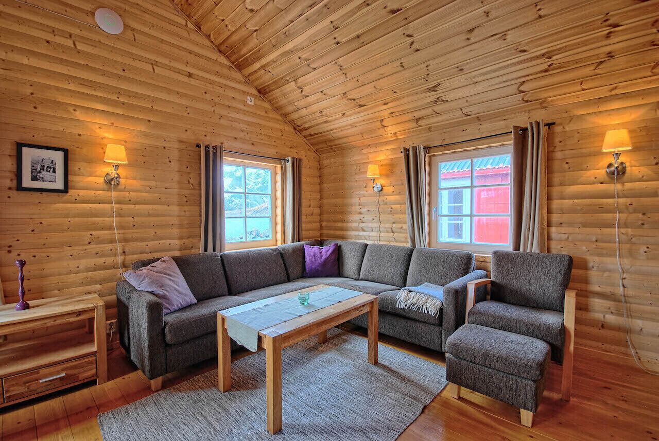 A cozy log cabin interior with a wooden ceiling, a sofa, a chair, a small table, and two wall-mounted lamps.