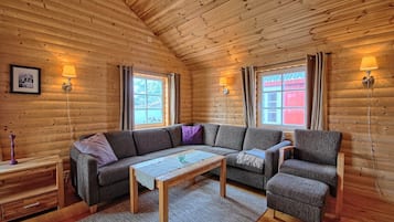 A cozy log cabin interior with a wooden ceiling, a sofa, a chair, a small table, and two wall-mounted lamps.