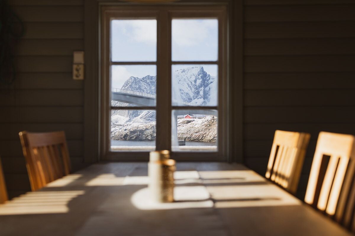 Un comedor con una mesa y sillas, una ventana con vistas a montañas nevadas y un puente, y paredes de madera.