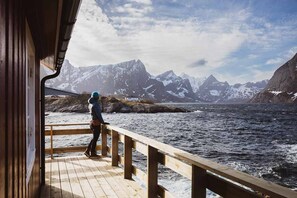 A wooden deck with a railing overlooking a fjord and snow-capped mountains.