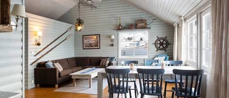 A cozy cabin interior with a wooden ceiling, a dining area with a table and chairs, a sofa, and a window with curtains.