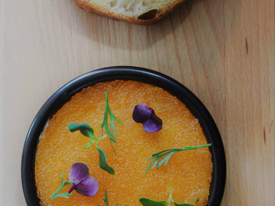 Un tazón de sopa de naranja decorado con hierbas y flores, acompañado de rebanadas de pan sobre una mesa de madera.