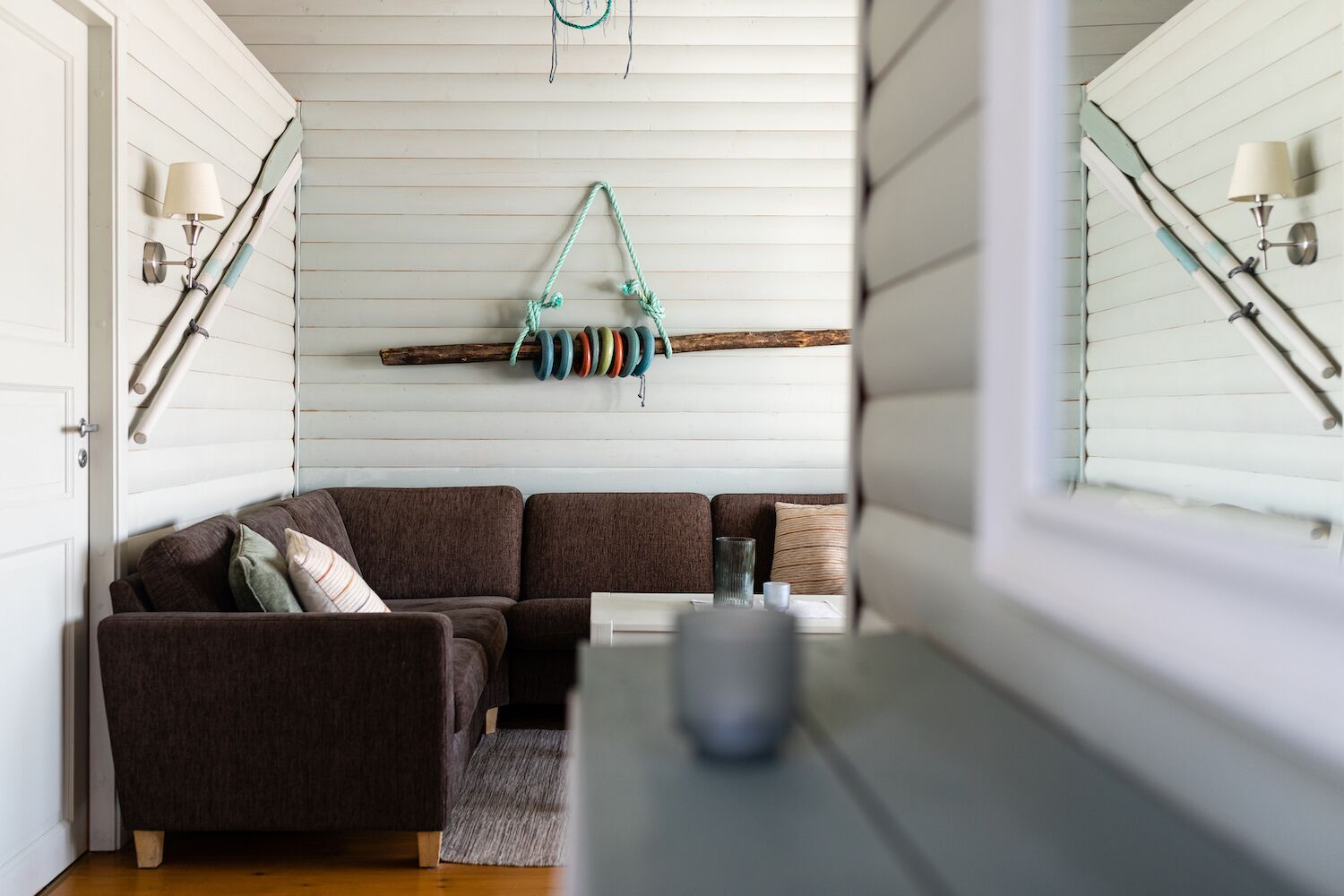 A cozy living room with a brown sectional sofa, a small rug, and a wooden coffee table. A colorful rope swing hangs from the ceiling, and a dreamcatcher is visible.