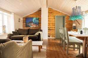 A cozy living room with a wooden ceiling, a painting of the aurora borealis, a dining table with chairs, and a sofa with cushions.