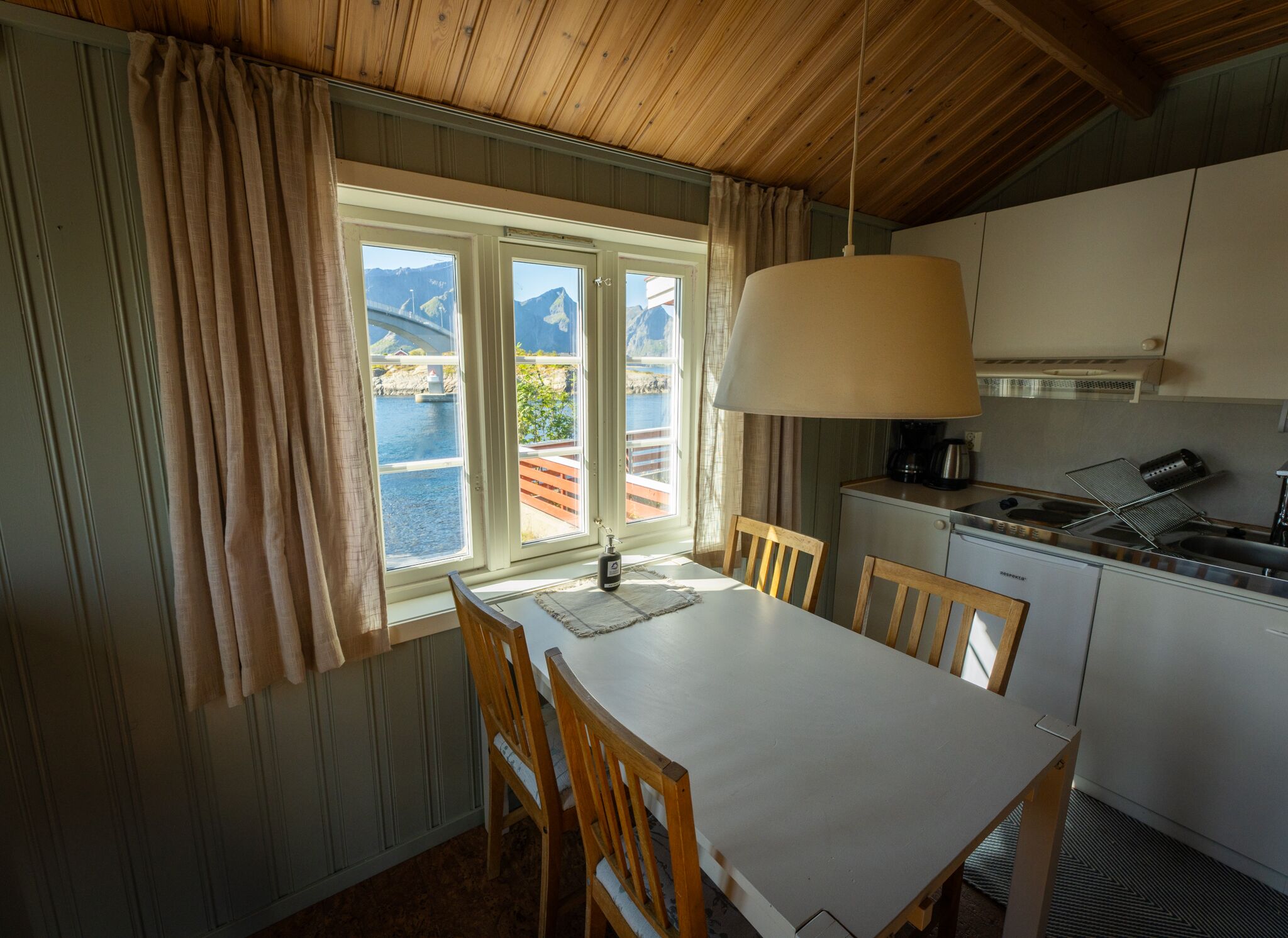 A kitchen with a dining table, chairs, a hanging lamp, and a view of a body of water through large windows.