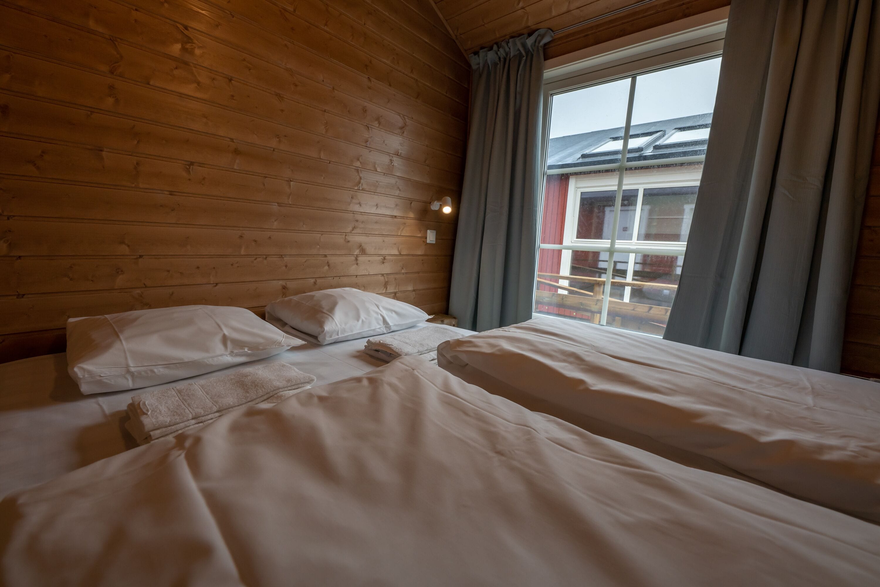 A wooden cabin room with two beds, a window with curtains, and a view of a building outside.