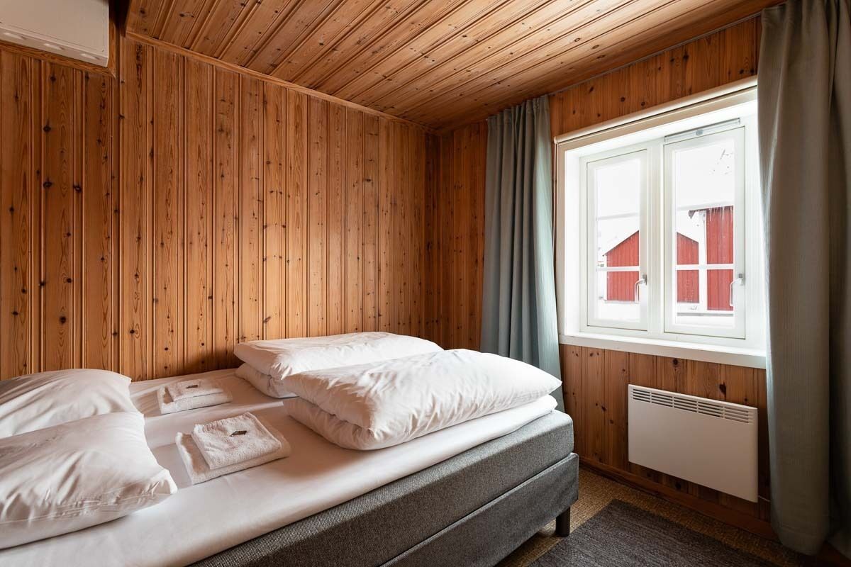Une pièce aux murs lambrissés, avec un lit, une fenêtre avec des rideaux et un radiateur.