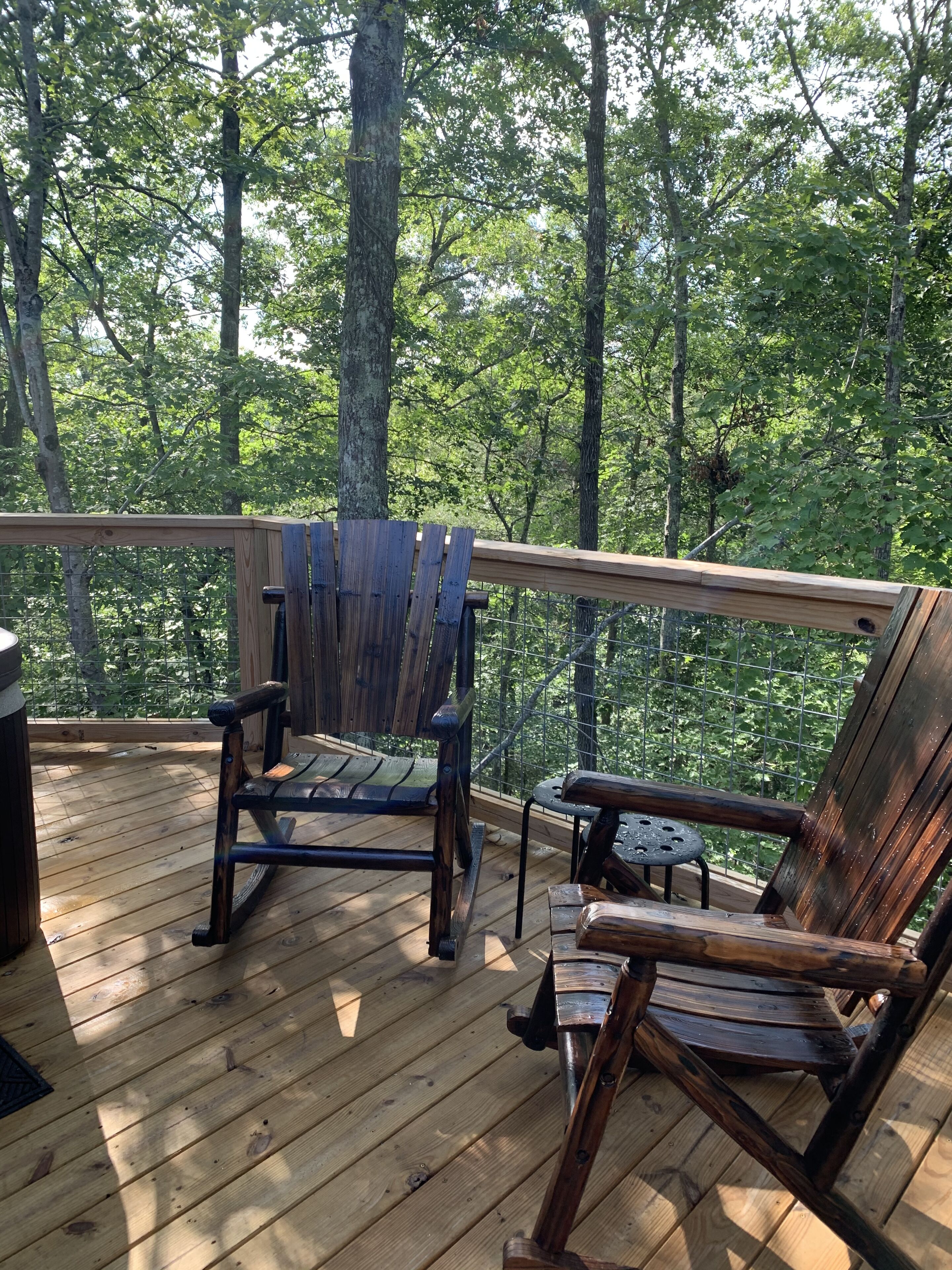 A wooden deck with two Adirondack chairs and a small table, surrounded by a metal fence and trees.