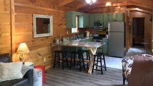 Un interior de cabaña acogedor con techo de madera, una cocina con armarios verdes, una zona de comedor con taburetes y una chimenea.