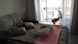 Ein ordentlich bezogenes Bett mit einer pinkfarbenen Decke, einem Blumenstrauß und Blick auf die Stadt durch große Fenster.