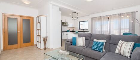 A modern living room with a grey sofa, glass coffee table, and a kitchen in the background.