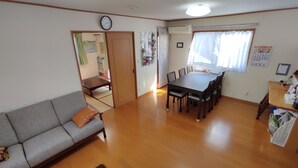 Ein Wohnzimmer mit einer Couch, einem Esstisch mit Stühlen, Holzboden, einer Uhr und einem Kalender an der Wand.
