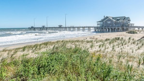 A beachfront property with a large house, a pier extending into the ocean, and a sandy beach with dunes in the foreground.