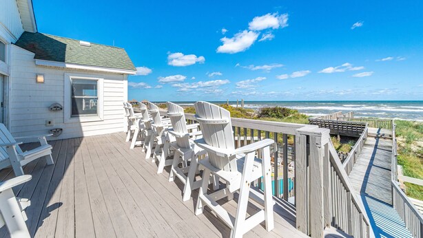A wooden deck with white chairs and a small building, overlooking a beach and ocean.