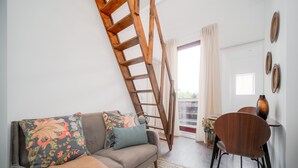 A cozy living room with a wooden ladder, a sofa with floral cushions, a dining table with chairs, and a view through a glass door. - Nomads Nest in Bonfim 1BED Balcony View (Porto)