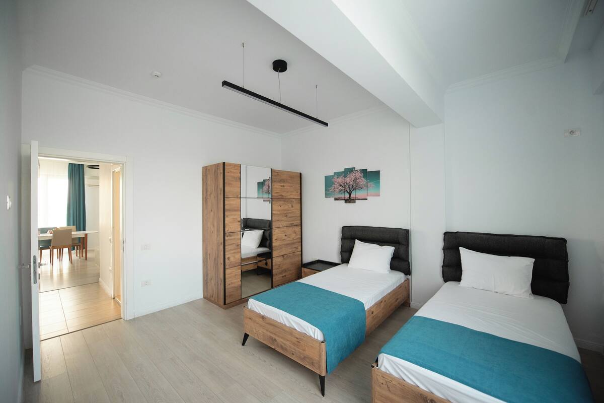 A modern bedroom with two beds, a wooden wardrobe, a desk, and a dining area.