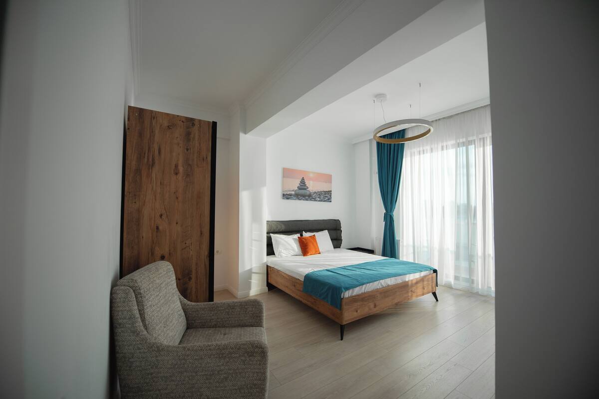 A modern bedroom with a wooden bed, a grey armchair, and a large window with sheer curtains.