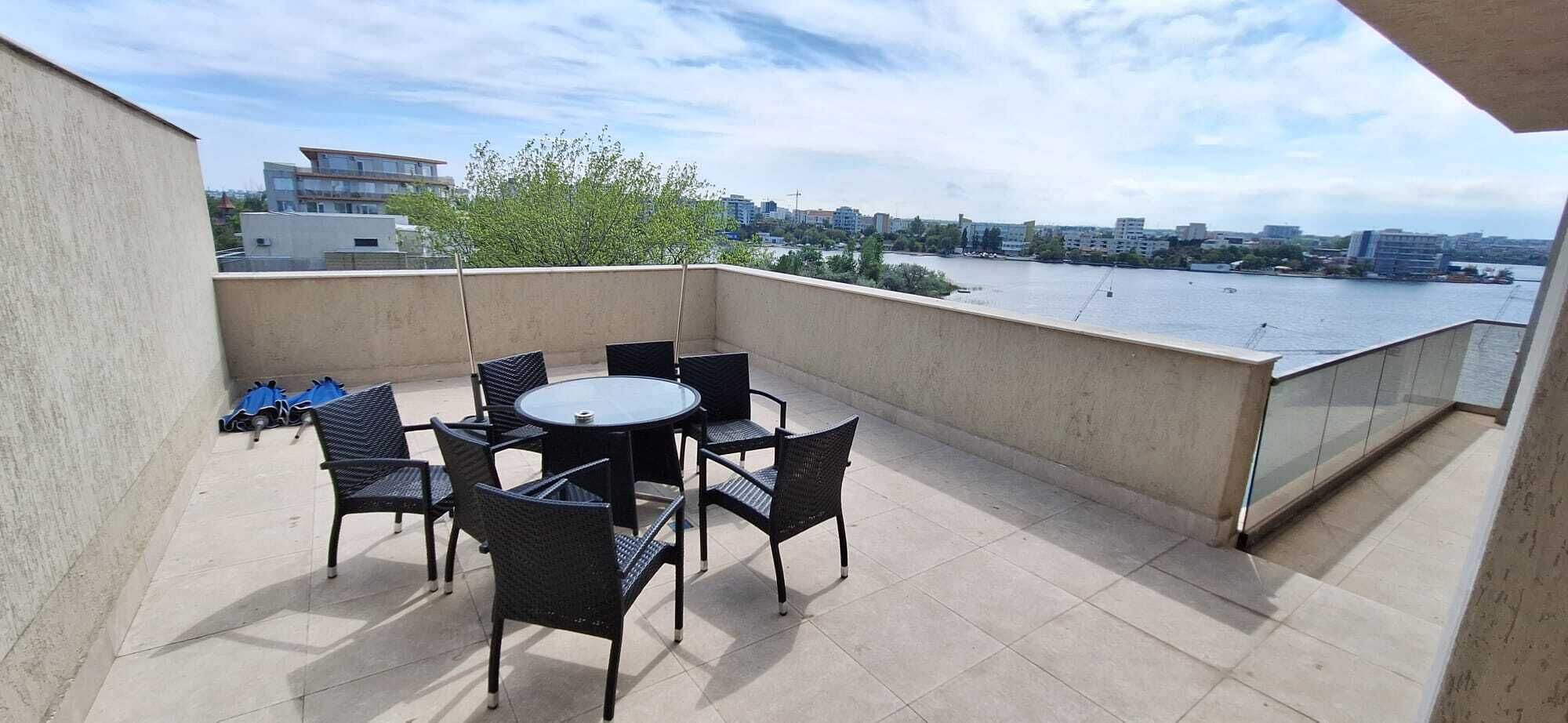A balcony with a round table and chairs, overlooking a body of water and cityscape.