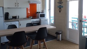 Cocina pequeña con armarios blancos, una mesa de madera y sillas negras. Incluye microondas, hervidor y un lavamanos.