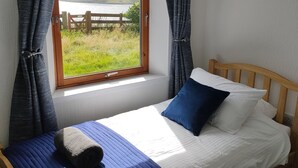 Een enkel bed met een houten hoofdbord, een raam met gordijnen en uitzicht op een gazon en een houten hek.