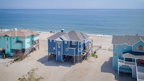 A beachfront property with multiple blue buildings, including a large central structure with a balcony and a smaller building with a red door. - 7048- Tide Song - Dog-Friendly Oceanfront Rodanthe Retreat (Rodanthe)