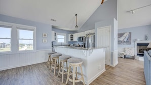 A kitchen with a bar area, wooden stools, a view through large windows, and a fireplace. - 7048- Tide Song - Dog-Friendly Oceanfront Rodanthe Retreat (Rodanthe)