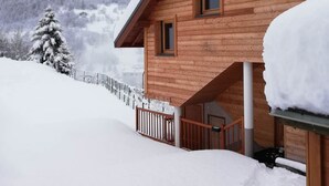 A wooden cabin with a covered porch and a snow-covered roof, surrounded by a snowy landscape and mountains. - Gîte 3- 10P- 4 bedrooms- Panoramic Balcony (VALMEINIER)