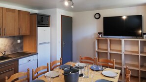 A compact kitchen with a microwave, refrigerator, and wooden cabinets. There is a dining table with chairs, a TV on a stand, and a clock on the wall. - Gîte 3- 10P- 4 bedrooms- Panoramic Balcony (VALMEINIER)