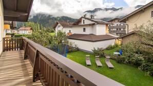 A balcony with a wooden railing overlooking a garden with sun loungers and a view of a mountain range. - Centrally located holiday home with a garden (Zell am Ziller)