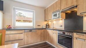 A modern kitchen with wooden cabinets, a built-in oven, a microwave, and a window with a view of a building. - Centrally located holiday home with a garden (Zell am Ziller)